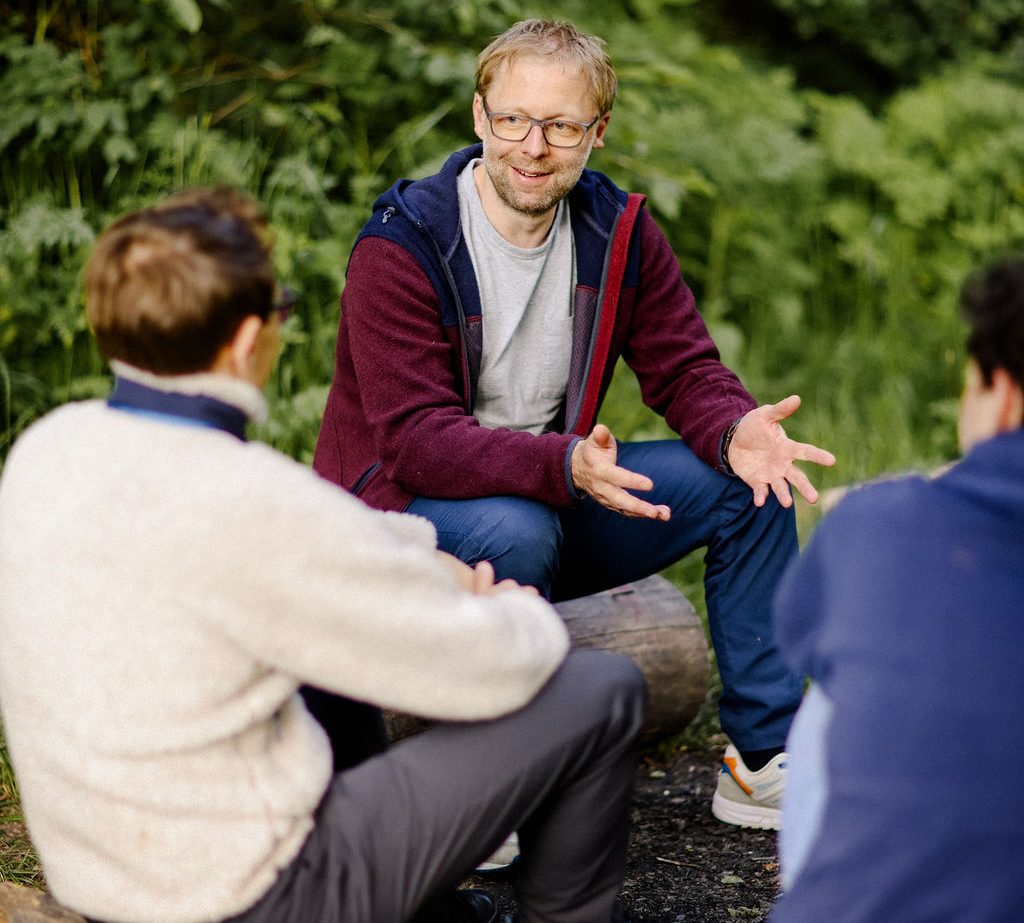 Gespräch Mann im Gespräch mit zwei Personen im Freien, umgeben von grüner Vegetation.
