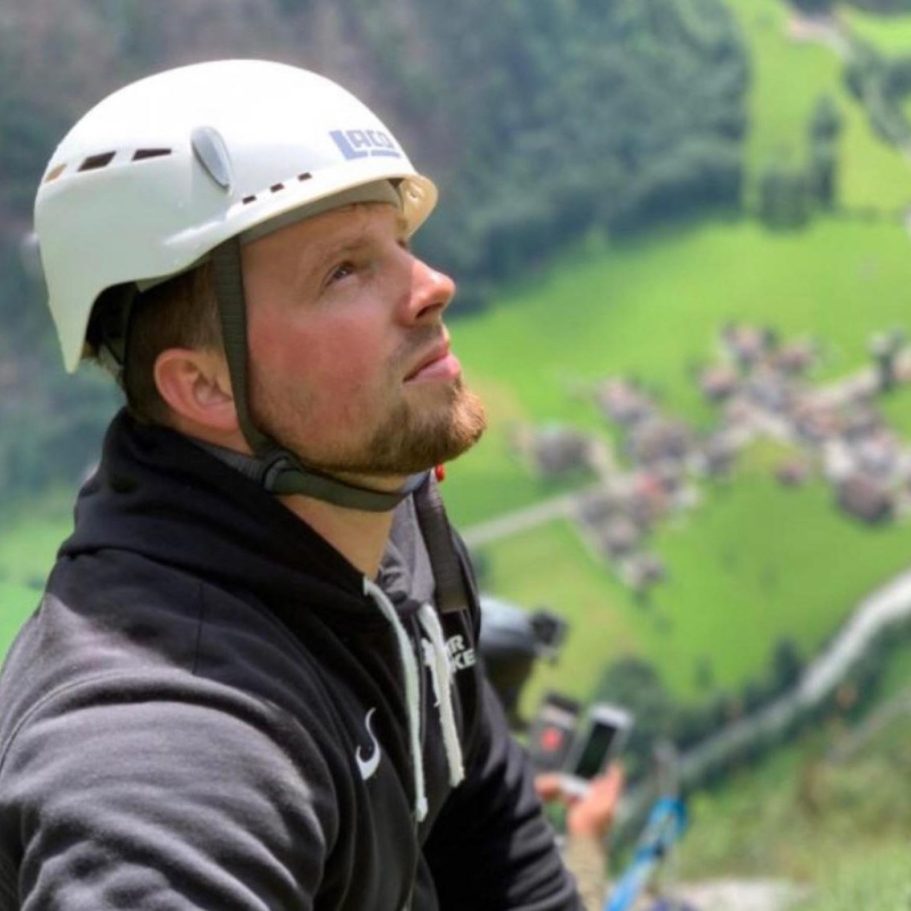 Coachee Mann mit Brille und Pullover, lächelt vor einer grünen Hintergrundlandschaft.