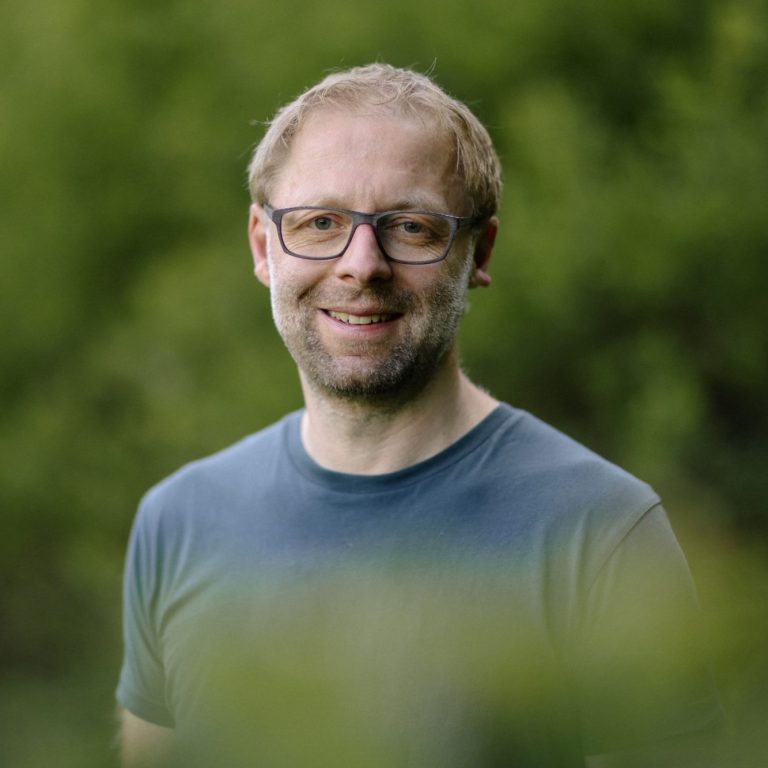 Coach Männlicher Porträt mit Brille, lächelnd vor unscharfem grünem Hintergrund.